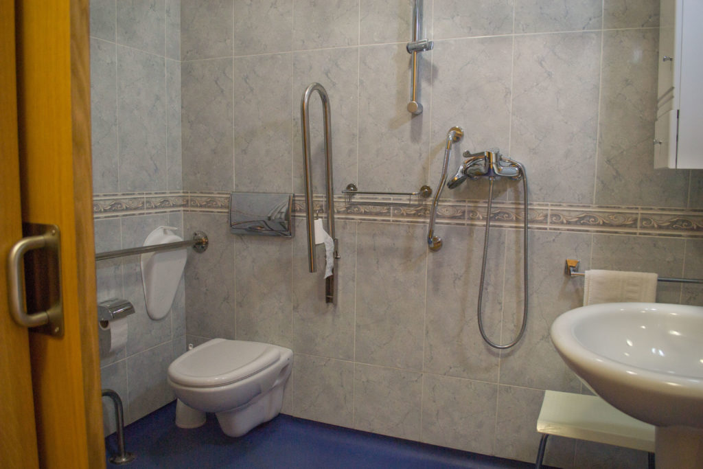 Baño geriátrico individual en residencia Ciudad de Aranda, con wc, ducha con asiento, armario y lavabo, con múltiples dispositivos de agarre y suelo antideslizante para comodidad y seguridad.
